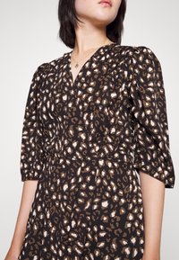 Leopardmönstrad klänning i brunt och krämfärgat, med V-ringning, puffärmar och figurnära midja. Tillverkad av lätt tyg med en mjuk textur.