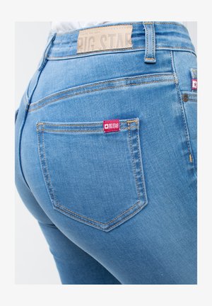 Kék farmernadrág, amely testhezálló kialakítással rendelkezik, a hátsó zseben egy kis piros címkével és kontrasztos varrással a varratok mentén.