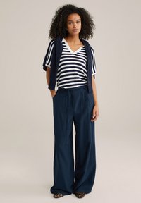 Tumed laiad püksid, mis on kombineeritud valge ja tumepunase triibu mustriga kootud ülevalt. Riietuse juurde kuulub ka tumepunane kardigan, mis on õlgadele visatud.