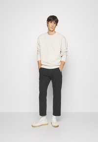 Lichtbeige sweatshirt met een textuurpatroon, donkere broek en witte sneakers met beige accenten. Model staat met de handen in de zakken.