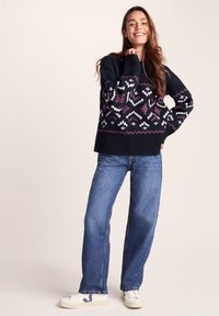 Smiling woman stands with hand on hip wearing black patterned sweater, blue jeans, and white sneakers against neutral background.