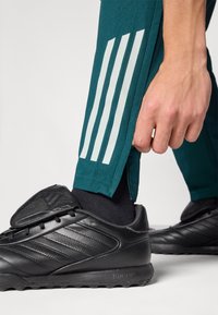 Teal athletic pants with three white vertical stripes on the lower leg, paired with a black sneaker featuring textured accents and lace-up design.