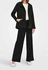 Černá kapucová bunda s kapsami, se zipovým uzávěrem a nastavovací šňůrkou. Spárována s volnými černými kalhotami a bílými botami.