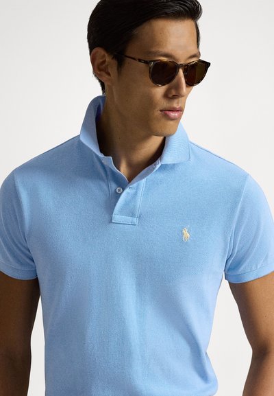 Γαλάζιο πόλο πουκάμισο από υφασμάτινη ύφανση με κλασικό γιακά, που διαθέτει μικρό κεντημένο λογότυπο σε χρυσό στο αριστερό στήθος.