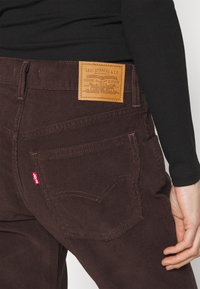 Pantalones de pana marrones con un parche de cuero que presenta el logo "Levi Strauss & Co.". Costuras distintivas y una pequeña etiqueta roja en el bolsillo izquierdo.