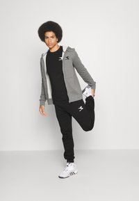 Harmaa vetoketjuhuppari mustan t-paidan päällä, mustat collegehousut ja valkoiset urheilukengät. Sisältää näkyvän logon vaatteessa.