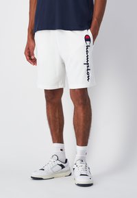 Pantalones cortos deportivos blancos con un logo "Champion" en azul marino en el lado izquierdo. Combinados con zapatillas blancas y calcetines blancos con acentos de logo.