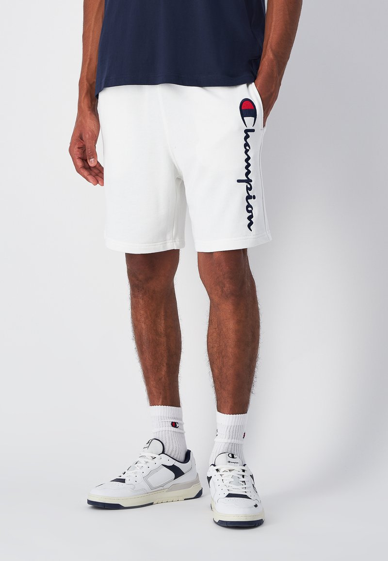 Pantalones cortos deportivos blancos con un logo "Champion" en azul marino en el lado izquierdo. Combinados con zapatillas blancas y calcetines blancos con acentos de logo.