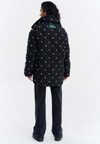 Czarna pikowana kurtka z różowymi kwiatowymi wzorami, wysokim kołnierzem i zieloną etykietą z tyłu. Luźny fason i kontrastujące ciemne spodnie.