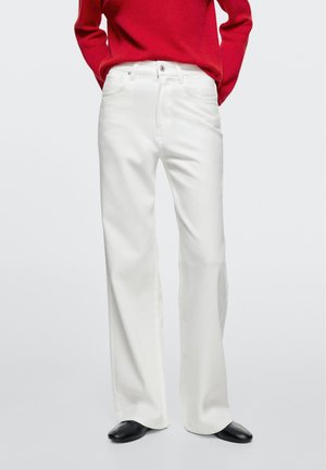 Personne portant un pantalon blanc taille haute et jambes larges, un haut rouge à manches longues rentré à l'intérieur, et des chaussures noires à bout pointu, debout devant un fond uni.