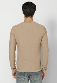 Beige, hosszú ujjú ing, ribbed textúrával, kerek nyakkal és testhez simuló kialakítással. Világos farmer nadrággal viselve, hátulról látható.