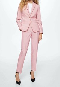 Jasnoróżowy szyty na miarę garnitur z dopasowaną marynarką i prostymi spodniami, z dwoma guzikami i bocznymi kieszeniami, w połączeniu z czarnymi szpilkami.