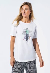 Weißes T-Shirt mit kurzen Ärmeln, das eine grafische Illustration einer Frau in einem grünen Oberteil und schwarzen Hosen zeigt, die mehrfarbige Einkaufstaschen hält.