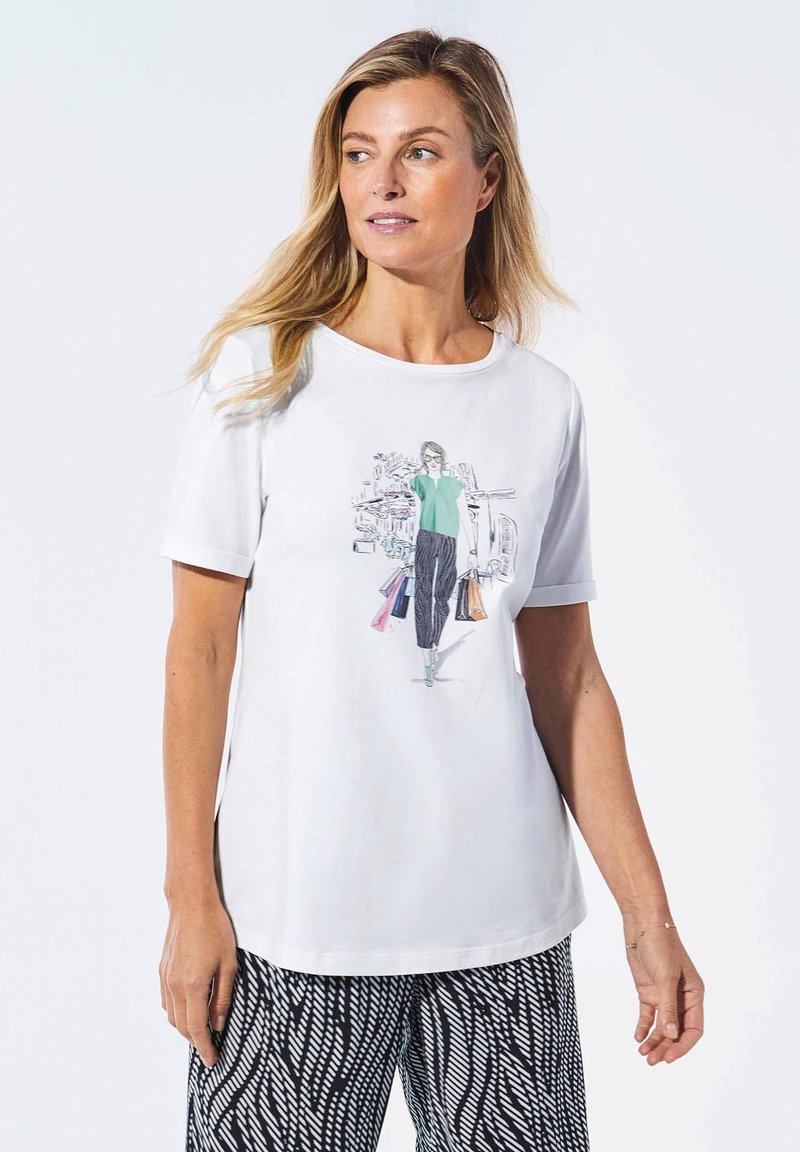 Weißes T-Shirt mit kurzen Ärmeln, das eine grafische Illustration einer Frau in einem grünen Oberteil und schwarzen Hosen zeigt, die mehrfarbige Einkaufstaschen hält.