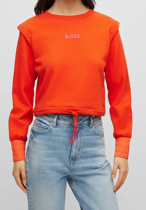 Nainen, jolla on päällään kirkkaan oranssi pitkähihainen lyhyt collegepaita, jossa on teksti "BOSS" ja kiristysnauhallinen helma, yhdistettynä vaaleansinisiiin korkeavyötäröisiin farkkuihin.