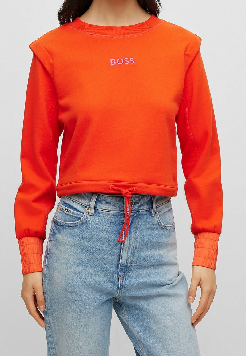 Femme portant un sweat-shirt court à manches longues orange vif avec l'inscription "BOSS" et un ourlet à cordon de serrage, associé à un jean taille haute bleu clair.