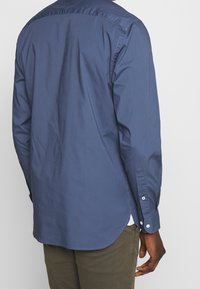 Mörkblå skjorta med knappar, med en slät textur, rundad nederkant och kontrasterande vita knappar på ärmarnas manschetter och knäppning. Avslappnad design.