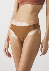 Bruine bikini-onderbroeken met een gladde textuur en zijaccenten. Huid toont vitiligo-vlekken; achtergrond is eenvoudig wit.