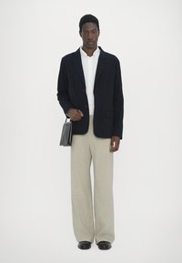 Black tailored blazer over a white shirt, paired with light beige wide-leg trousers. Holds a small black handbag; black shoes complete the outfit.