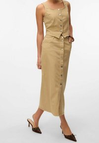 Vestido de mezclilla caqui con botones, parte superior sin mangas y corta, falda larga, bolsillos delanteros y botones plateados. Llevado con mules de tacón marrones.