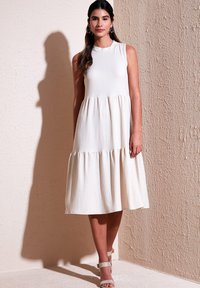 Sleeveless cream dress with a fitted top and tiered, flowing hem. Made of textured fabric, featuring a scoop neckline and subtle ripples.