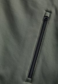 Olivgrön tyg med en zippered ficka med svarta detaljer. Texturen är jämn, och designen är funktionell med en elegant finish.