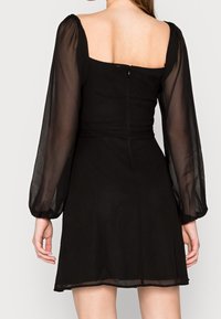 Svart klänning med en åtsittande bolero med fyrkantig rygg, genomskinliga puffärmar och en utställd kjol. Osynlig dragkedja på ryggen.