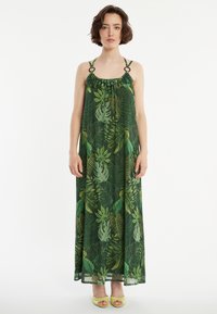 Maxikleid mit grünen Blattmustern, leichtem Stoff, rüschigem Halsausschnitt und großen Ringen an den Schulterträgern. Getragen mit gelben Absätzen.