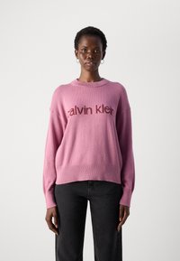 Maglione rosa lavorato a maglia con il logo "calvin klein" in una tonalità più scura, maniche larghe e orlo a costine, abbinato a pantaloni neri.