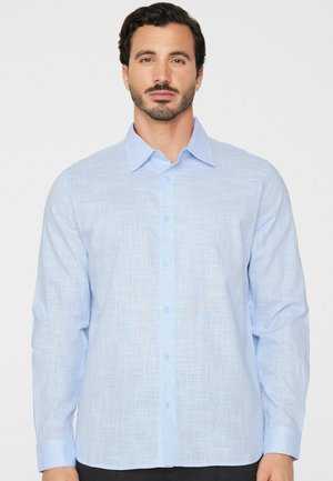 Mann mit dunklem Haar und Bart, der ein hellblaues, strukturiertes Langarmhemd mit Knöpfen trägt und auf einem schlichten Hintergrund nach vorne blickt.