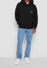 Sudadera con capucha negra con el logo azul de "Calvin Klein Jeans" en el pecho, combinada con vaqueros rectos de color azul claro y zapatillas blancas.