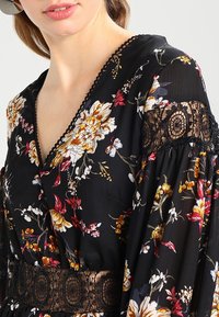 Blouse noire à motifs floraux avec des manches longues, présentant des imprimés floraux colorés, un col en V et des détails en dentelle autour des manches et de la taille.
