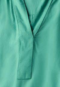 Turkise bluse med V-hals og struktureret stof. Har ren syning og et enkelt foldet panel som accent nær halsen.