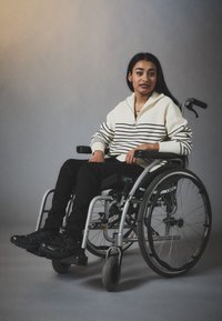 Una sedia a rotelle argento e nera con ruote a raggi, dotata di un sedile intrecciato. L'utente indossa un maglione a righe bianche e nere e pantaloni neri.