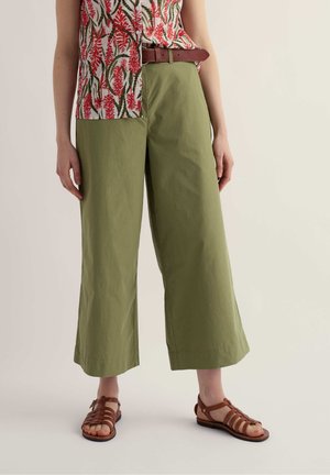 Frau trägt olivgrüne, weit geschnittene, knapp knöchellange Hose, braunen Ledergürtel, braune Sandalen mit Riemchen und eine ärmellose rote Bluse mit Blumenmuster.