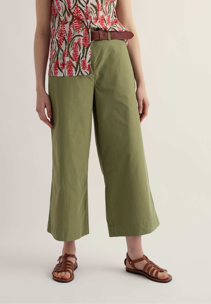 Femme portant un pantalon large coupé olive, une ceinture en cuir marron, des sandales marron à lanières, et un chemisier sans manches à fleurs rouges.