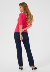 Tajt rosa topp med trekvarts ärmar, kombinerad med mörkblå jeans. Modellen har långt hår och står mot en neutral bakgrund.