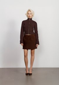 Suéter castanho canelado com fecho, de gola alta, emparelhado com uma saia mini castanha. Ambas as peças apresentam texturas suaves e cortes ajustados.