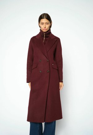 Donna in piedi rivolta in avanti, indossa un lungo cappotto doppio petto bordeaux sopra un dolcevita abbinato e jeans blu su sfondo bianco.