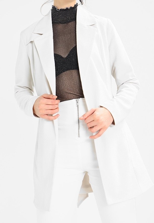 Biały długi żakiet z kołnierzem szalowym, noszony na przezroczystej czarnej bluzce. Biały materiał wydaje się gładki, a kroj cechuje się czystymi liniami.