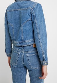 Džínová bunda s krátkým střihem, světle modré barvy, klasickým límcem a předními náprsními kapsami. Má knoflíkové manžety a jemné detaily v prošívání.