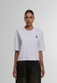 Weißes, kurzes Baumwoll-T-Shirt mit kurzen Ärmeln, das ein schwarzes Logo auf der linken Brust trägt. Glatte Textur mit Rundhalsausschnitt.