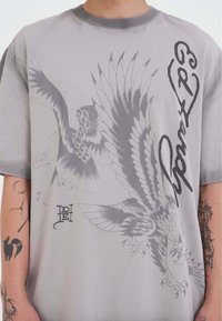 Camiseta de algodón gris con gráficos grandes de un águila gris y texto negro. Corte relajado con mangas cortas, escote redondo y estampados prominentes.