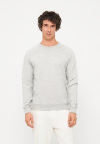 Jeune homme aux cheveux bouclés et foncés portant un sweat-shirt gris clair à manches longues et un pantalon blanc, debout devant un fond blanc uni.