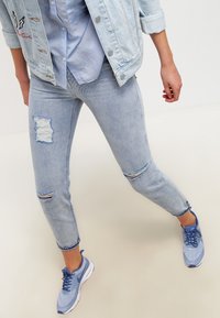 Henkilö, joka käyttää vaaleansinisiä revittyjä farkkuja, farkkutakkia ja sinisiä Nike-kenkiä, seisoo jalat harallaan tasaisella taustalla.