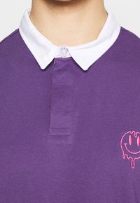 Kragen und oberer Brustbereich einer Person, die ein lila Polo-Shirt mit weißem Kragen und einer pinken, tropfenden Smiley-Gesicht-Stickerei auf der rechten Seite trägt.
