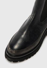 Botte en cuir noir avec un design élégant, des côtés élastiques et un accent métallique texturé près de l'orteil. Semelle en caoutchouc épaisse.