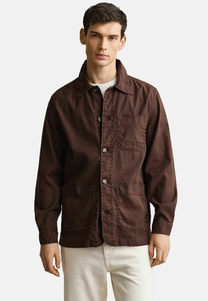 Jonge man met een bruin jasje met knopen en drie voorzakken, over een wit overhemd en lichtgekleurde broek, tegen een effen achtergrond.