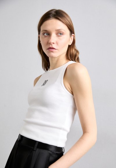 Weißes Baumwoll-Tanktop mit körperbetontem Schnitt, versehen mit einem kleinen schwarzen Logo auf der Brust, kombiniert mit einem schwarzen, plissierten Rock.