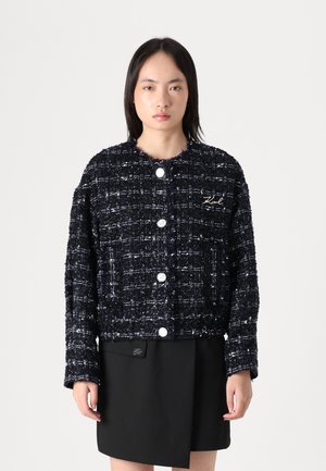Marineblauwe tweed jacket met een boxy ontwerp, witte accenten, ronde knopen en een subtiele logo-detail, gecombineerd met een zwarte rok.
